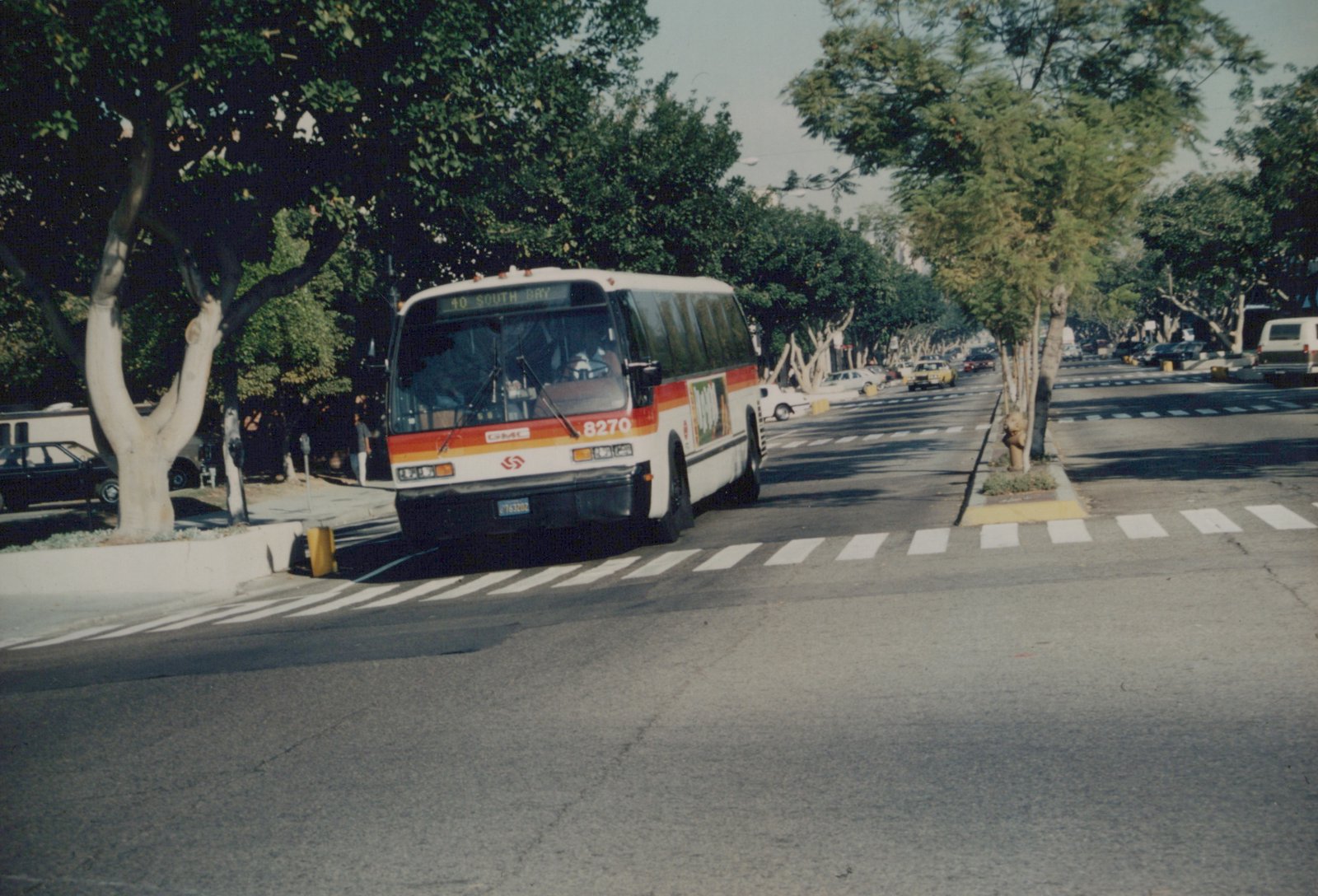 A white and red GMC bus numbered 8270, operated by Southern California Rapid Transit District, is turning on a tree-lined street in Los Angeles with a yellow taxi and other cars in the background.