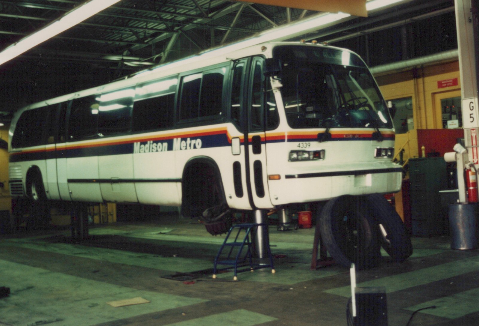 A white Madison Metro bus, number 4339, is elevated on a maintenance lift inside a garage with tires and tools nearby.