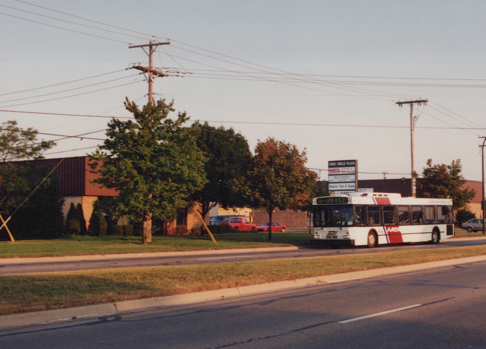 A white and red AATA bus with the number 308 is driving down a street, with trees and a shopping center sign in the background during dusk.