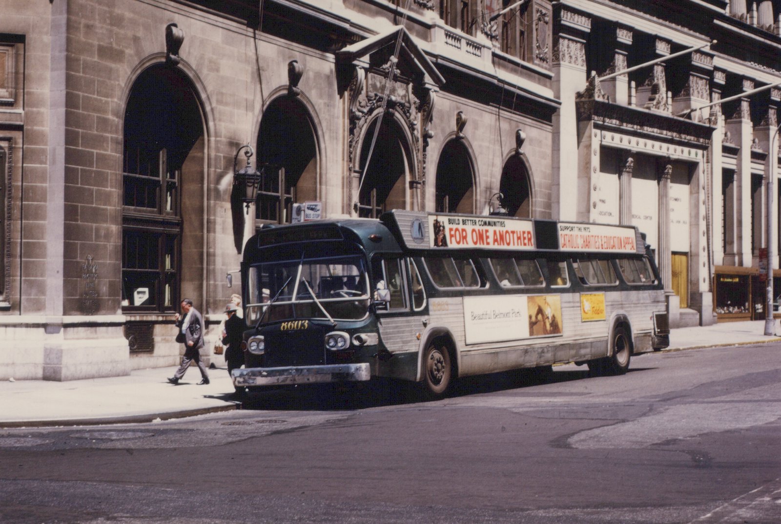 A dark green and silver city bus, numbered 8603, is parked on a street in front of a large stone building; a sign on its roof promotes the 1972 Catholic Charities appeal, and pedestrians walk on the sidewalk.