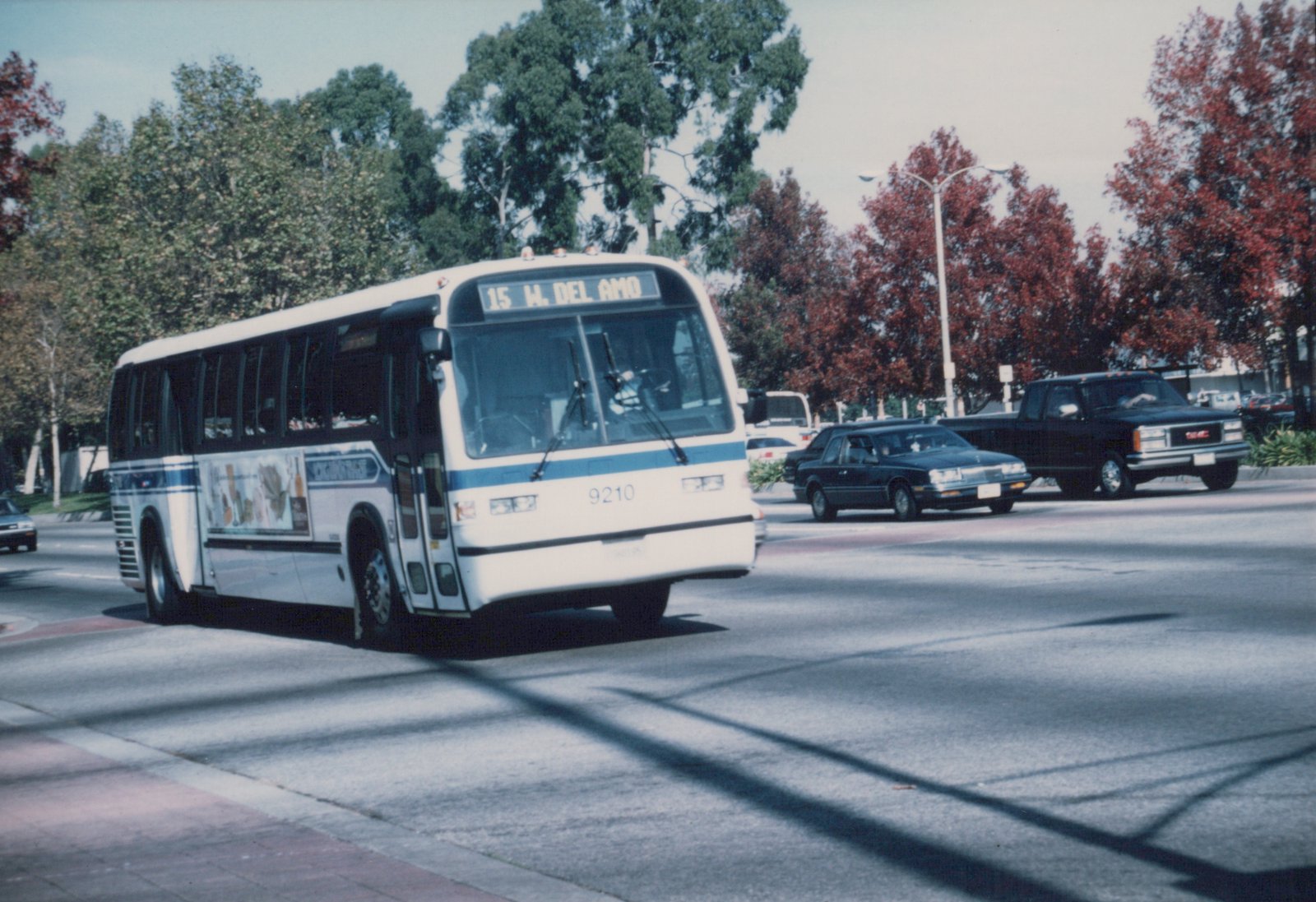 A white and blue motor bus with fleet number 9210 is driving on a city street, its destination sign reading '15 W. DEL AMO', with trees and other cars in the background.