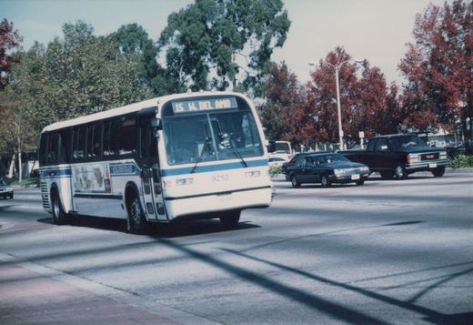 A white and blue motor bus with fleet number 9210 is driving on a city street, its destination sign reading '15 W. DEL AMO', with trees and other cars in the background.