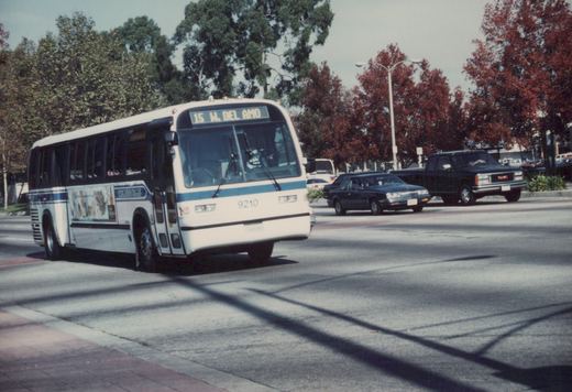 A white and blue SCRTD bus with the number 9210 is driving down a street with other cars and trees in the background.