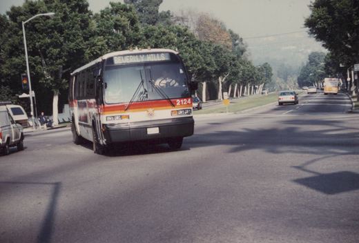 A white, red, and orange bus with number 2124 and a destination sign reading 'BEVERLY HILLS' is driving down a street lined with trees and other vehicles.