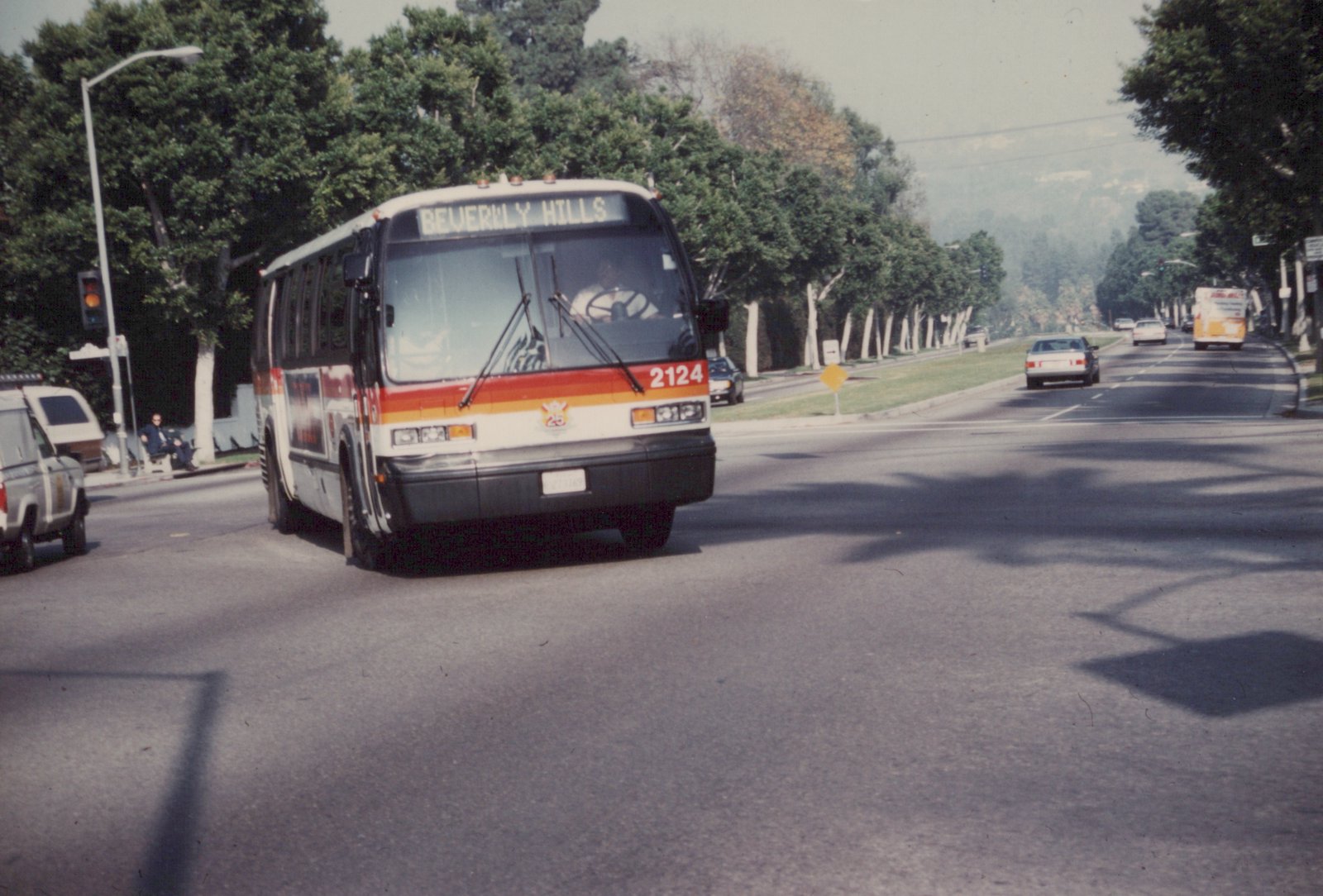 A white, red, and orange bus with number 2124 and a destination sign reading 'BEVERLY HILLS' is driving down a street lined with trees and other vehicles.