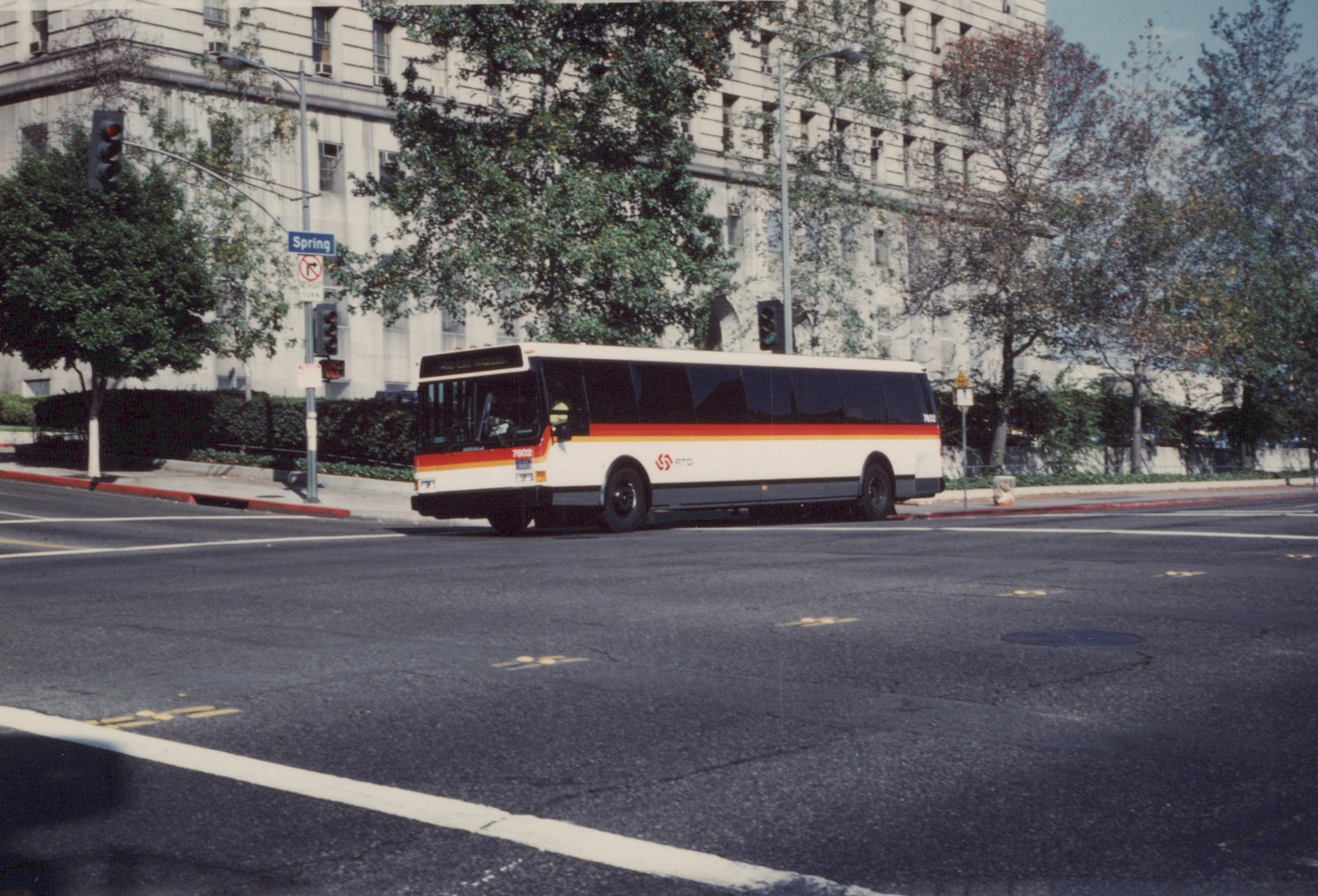 A white RTD bus with red and orange stripes, numbered 7602, is stopped at a street intersection in Los Angeles with a street sign reading 'Spring' visible in the background.
