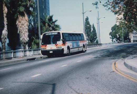 A white Metro bus with red and blue stripes, number 2474, is shown driving on a curved city street with palm trees and a glass skyscraper in the background.