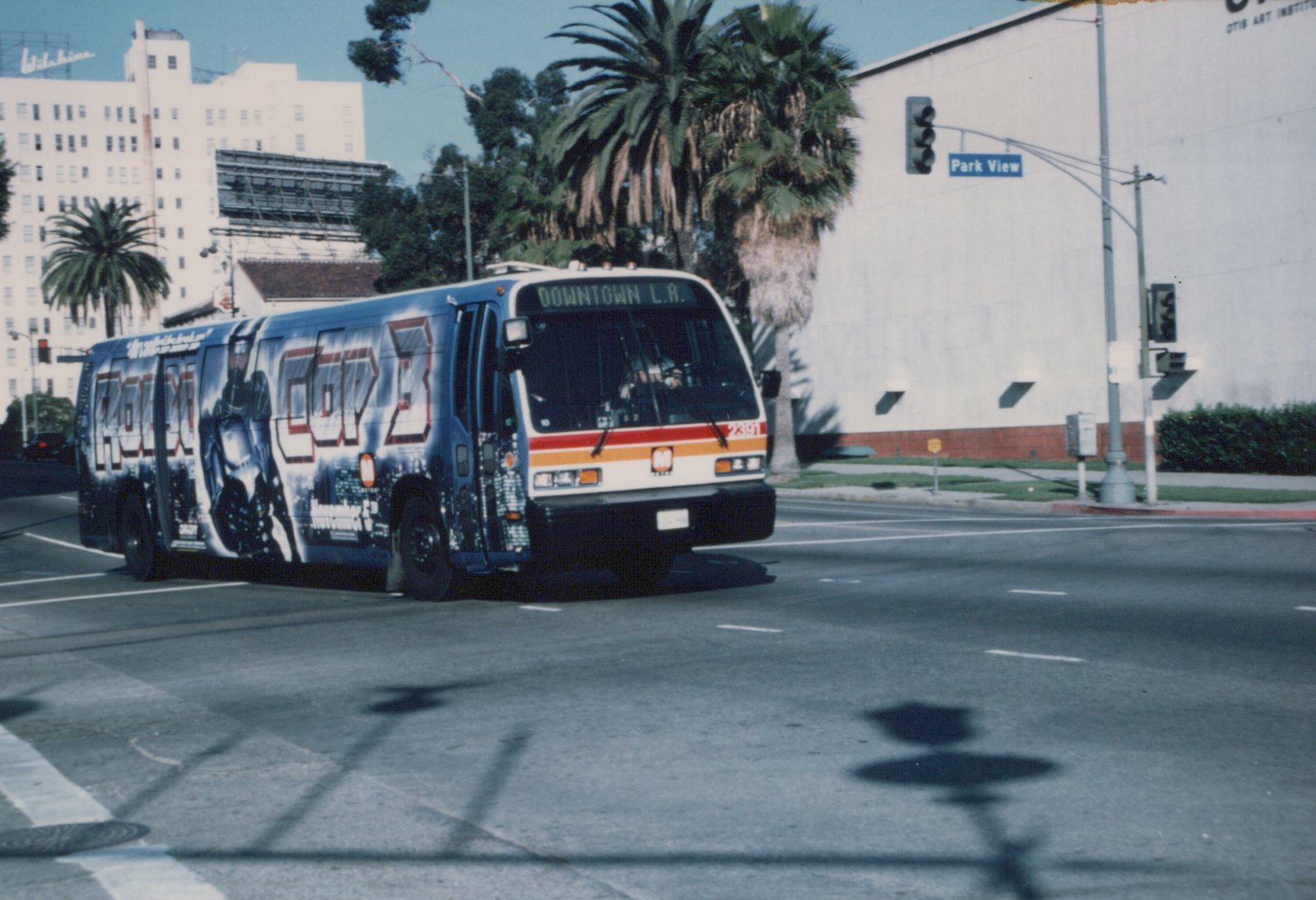 A city bus with a 'RoboCop 3' movie advertisement on its side is driving down a street with palm trees and a building in the background.
