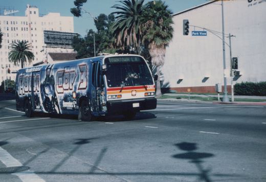 A city bus with a 'RoboCop 3' movie advertisement on its side is driving down a street with palm trees and a building in the background.