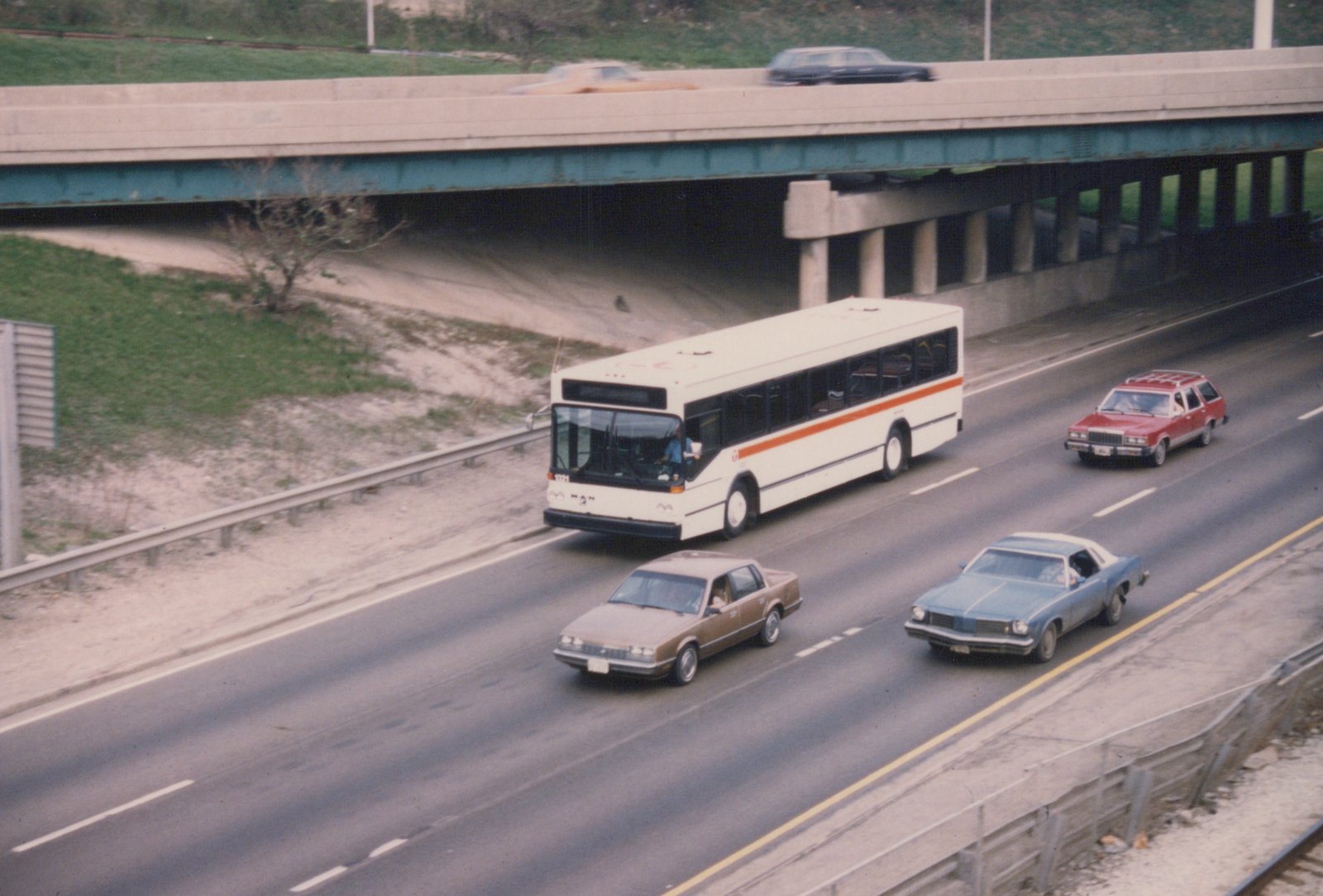A white and orange CTA bus drives on a multi-lane highway under an overpass, with several cars from the 1970s around it, including a red station wagon and blue coupe.