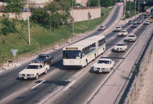 An STA bus drives down an expressway with other cars under a clear sky. The bus is white with blue and yellow stripes, and the number 653 is visible on its front.