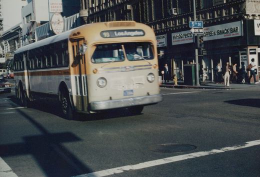 A vintage motor bus with a tan and silver livery, displaying route 93 to Los Angeles, is turning on a city street with pedestrians and storefronts including 'Whoopee's' visible in the background.