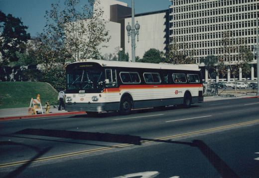 A white RTD bus with red and orange stripes, number 6111, is driving down a city street with modern buildings and trees in the background.