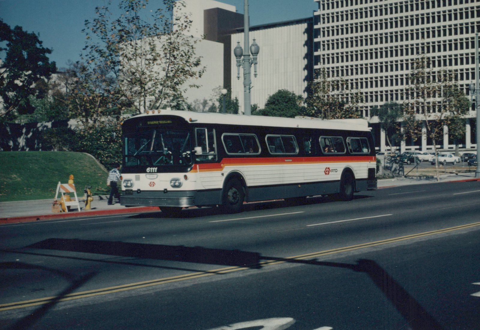 A white RTD bus with red and orange stripes, number 6111, is driving down a city street with modern buildings and trees in the background.