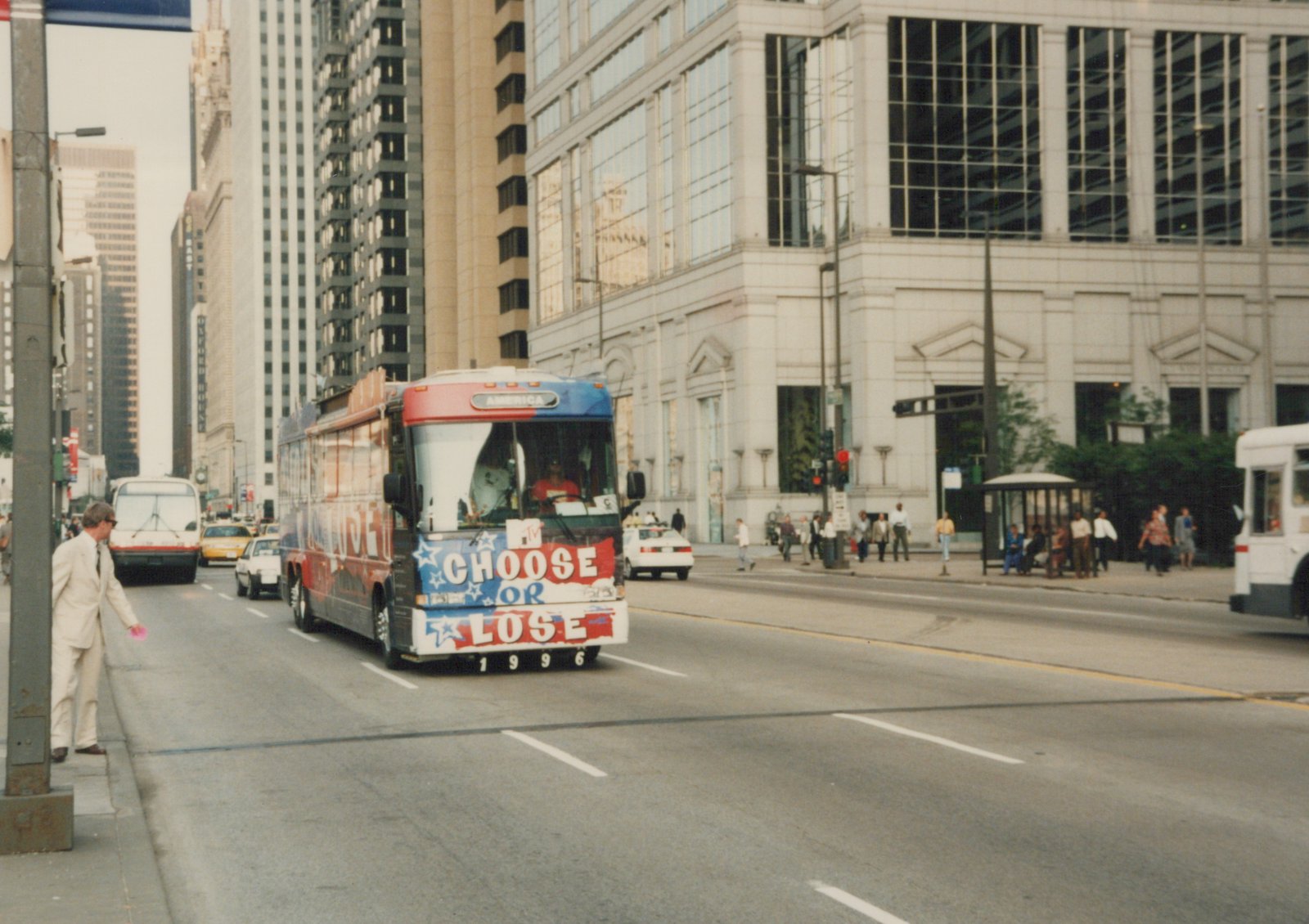 A red, white and blue MTV tour bus with 'CHOOSE OR LOSE 1996' on its front bumper travels down a city street lined with tall buildings and pedestrians.