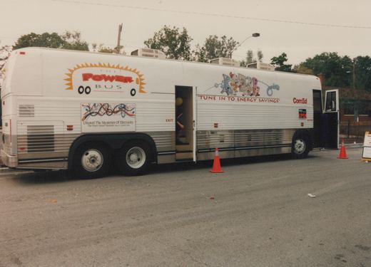 A white bus with 'THE POWER BUS' and 'ComEd' lettering, featuring cartoon characters and the slogan 'TUNE IN TO ENERGY SAVINGS', is parked on a street with its doors open and orange cones placed nearby, indicating it is set up for public display.