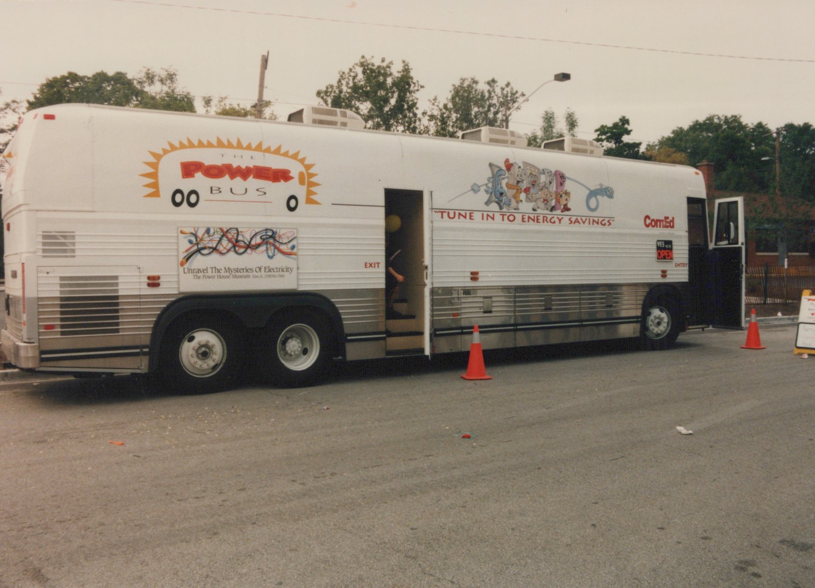 A white bus with 'THE POWER BUS' and 'ComEd' lettering, featuring cartoon characters and the slogan 'TUNE IN TO ENERGY SAVINGS', is parked on a street with its doors open and orange cones placed nearby, indicating it is set up for public display.