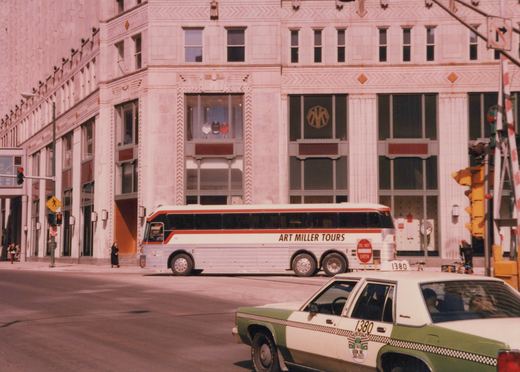 A white and red motor bus labeled 'Art Miller Tours' and numbered 1380 is parked on a city street beside a green and white Checker taxi. A large Art Deco-style building stands behind them.