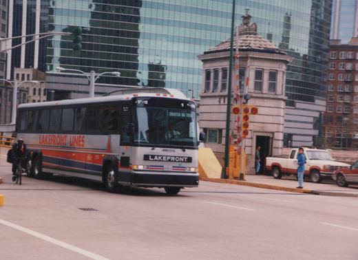 A silver Lakefront Lines motor bus with red and blue stripes, marked E-12 and 'CHARTER ON TOUR', drives down a city street past a stone bridge tender's house and glass skyscrapers.