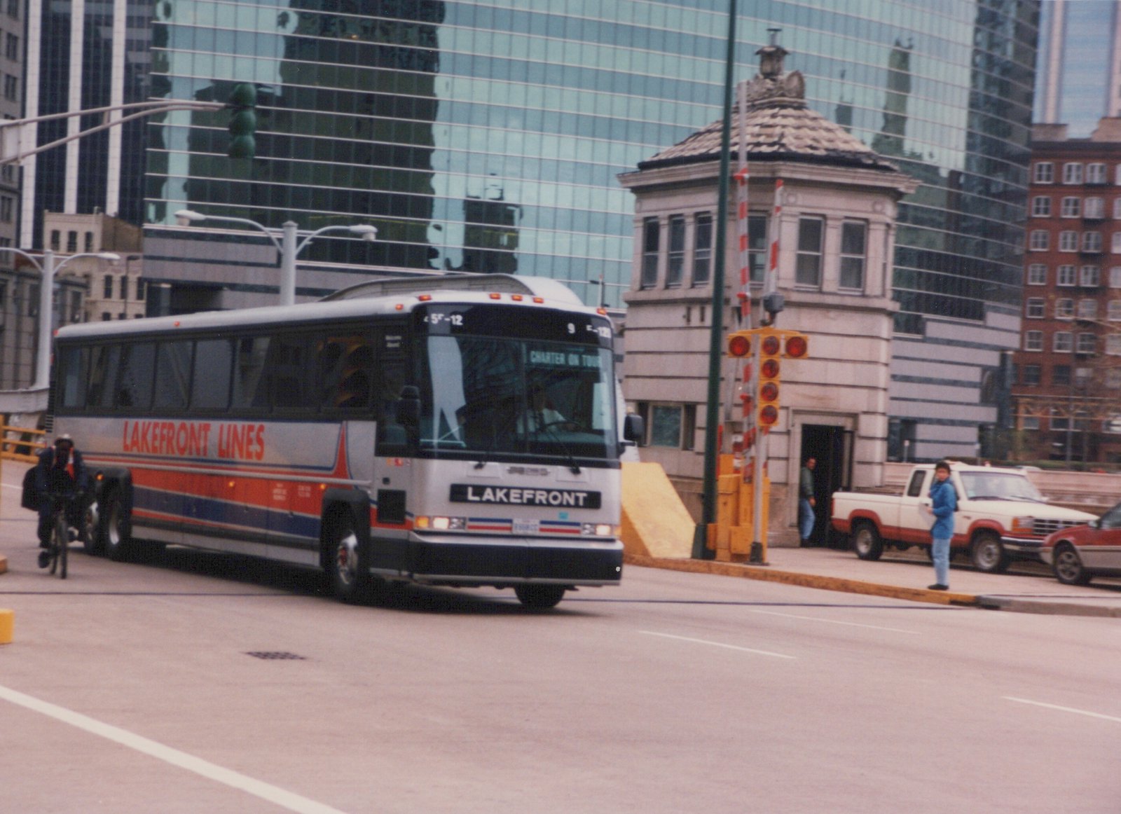 A silver Lakefront Lines motor bus with red and blue stripes, marked E-12 and 'CHARTER ON TOUR', drives down a city street past a stone bridge tender's house and glass skyscrapers.