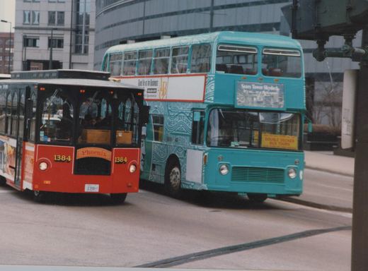 A red trolleybus, numbered 1384 and labeled 'Phoenix,' drives on a city street next to a turquoise double-decker tour bus. The scene features modern buildings and a visible overhead wire.