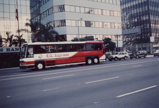 A side view of a red and white K.C. Express bus number 802 driving on a multi-lane city street with office buildings and other cars in the background.