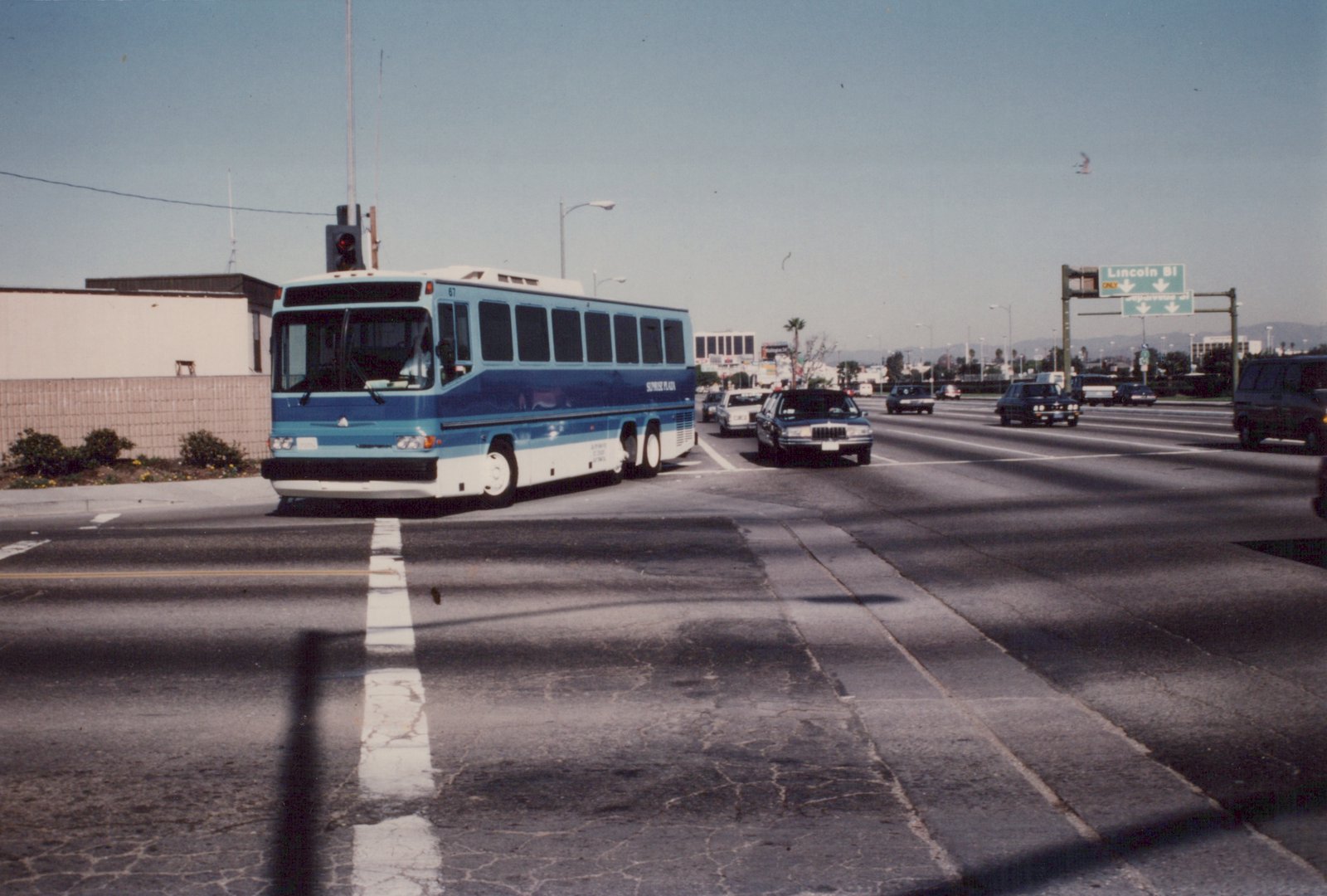 A blue and white motor bus labeled 'Sunrise Plaza' and numbered 67 is stopped at a street intersection during the day, with cars and a highway sign visible in the background.