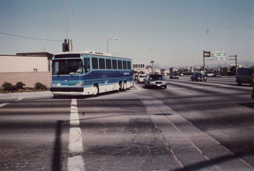 A blue and white motor bus labeled 'Sunrise Plaza' and numbered 67 is stopped at a street intersection during the day, with cars and a highway sign visible in the background.