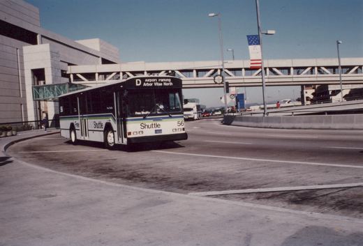 A white and green airport shuttle bus with the number 56 on its side and front, labeled 'Free LAX Shuttle' and 'Shuttle', is turning in a paved area near an airport terminal building with an elevated walkway in the background.