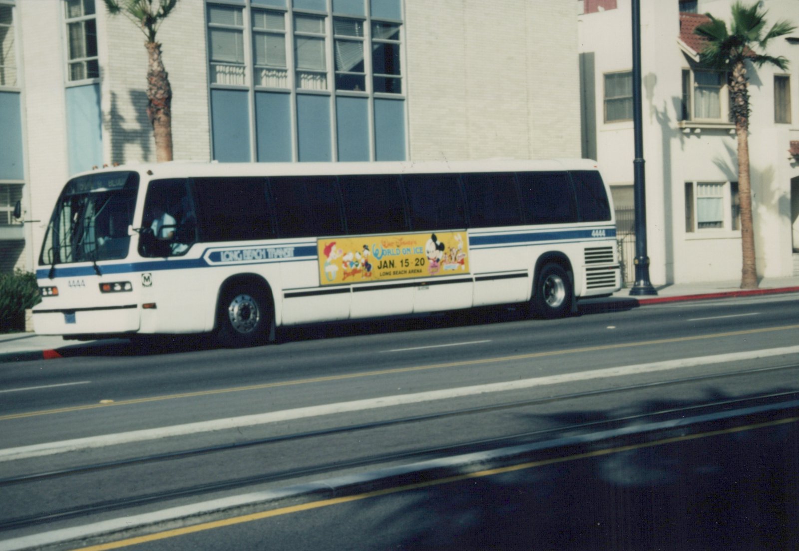 A white and blue transit bus with the number 4444 is stopped on a city street. The side of the bus has a large yellow advertisement for Walt Disney's World on Ice. Palm trees and buildings are visible in the background.