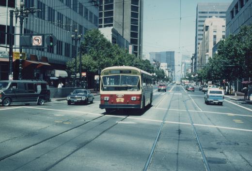 A red and cream trolleybus with the number 4009 on its front travels down a wide city street with visible trolley wires, surrounded by other cars and tall buildings on a sunny day.