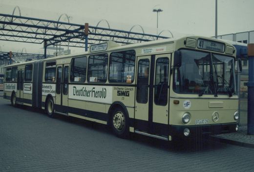 A cream and black articulated bus with 'Stadtwerke Gütersloh SWG 29' on its side and 'Deutscher Herold' advertising, parked under a blue metal canopy.