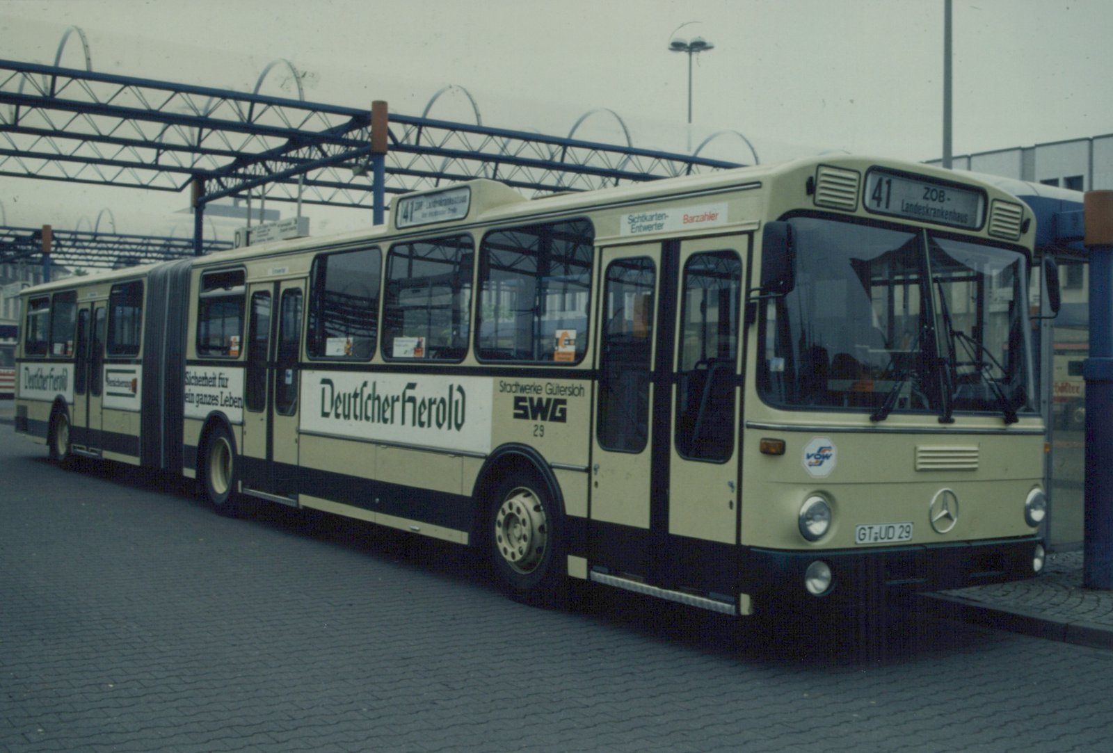 A cream and black articulated bus with 'Stadtwerke Gütersloh SWG 29' on its side and 'Deutscher Herold' advertising, parked under a blue metal canopy.