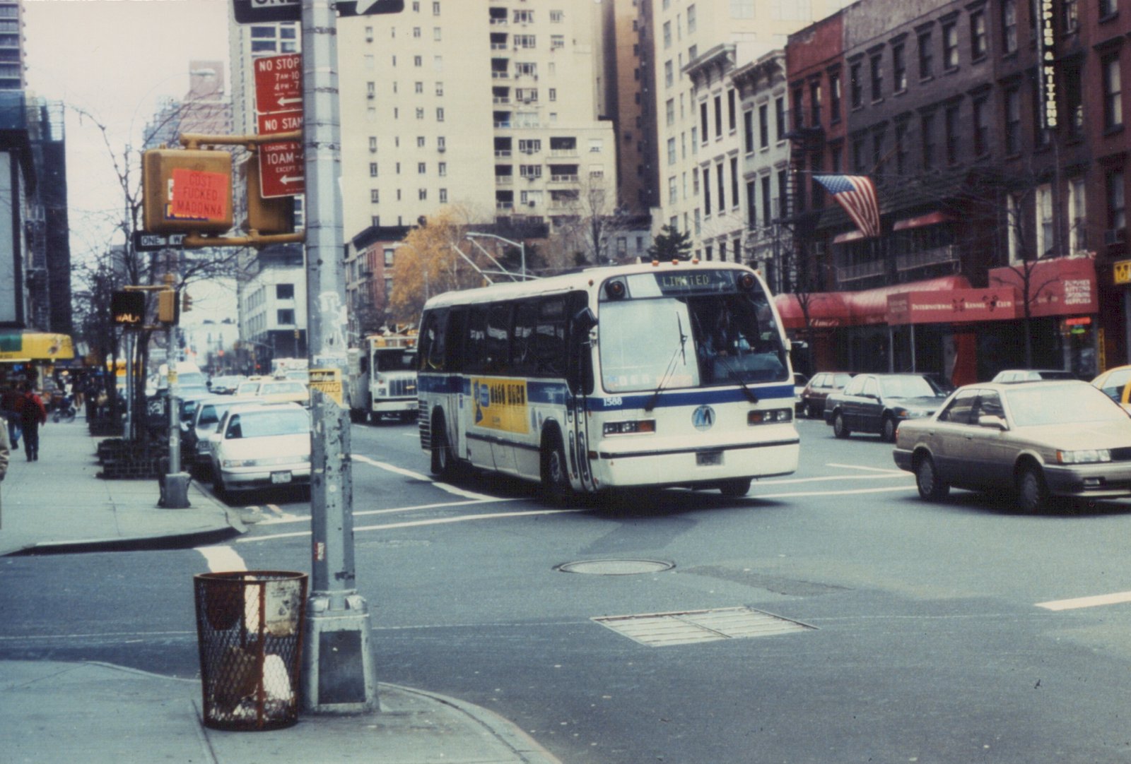A white and blue MTA bus with the number 1588 drives on a city street, passing by storefronts and parked cars under an overcast sky.