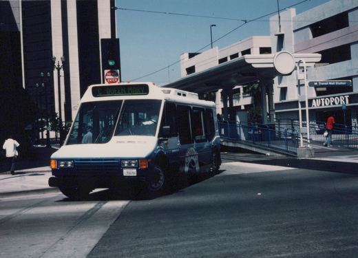 A white and blue Flxible Metro bus with the destination 'C QUEEN MARY' is stopped at the M Transit Mall Station, with a modern building and blue railings in the background.
