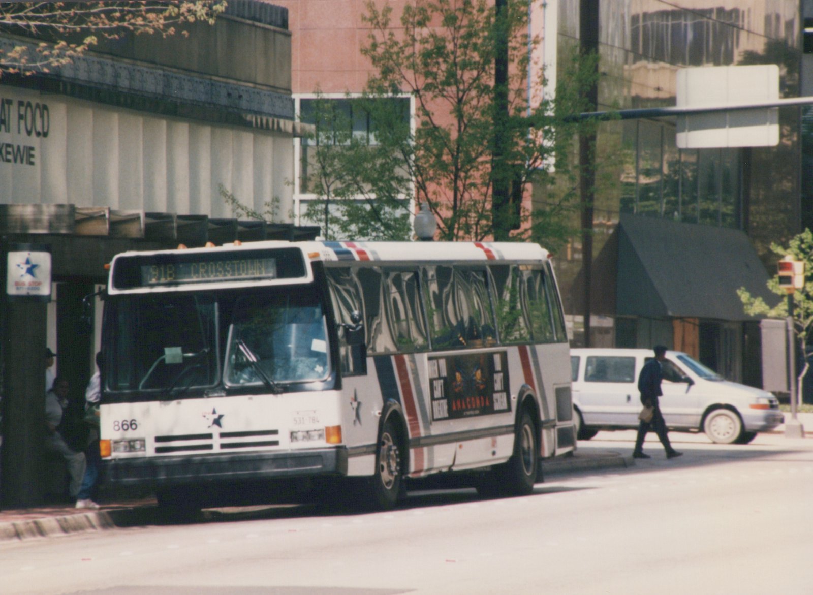 A white New Flyer D40 bus with red and blue stripes, number 866, is stopped at a city street corner with people nearby; the destination sign reads '91B CROSSTOWN'.