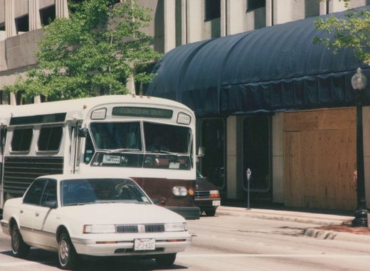 A white and brown Flxible Metro bus with 'COWTOWN BUS' on its sign is parked on a city street, with a white sedan in front and a building with a blue awning behind.