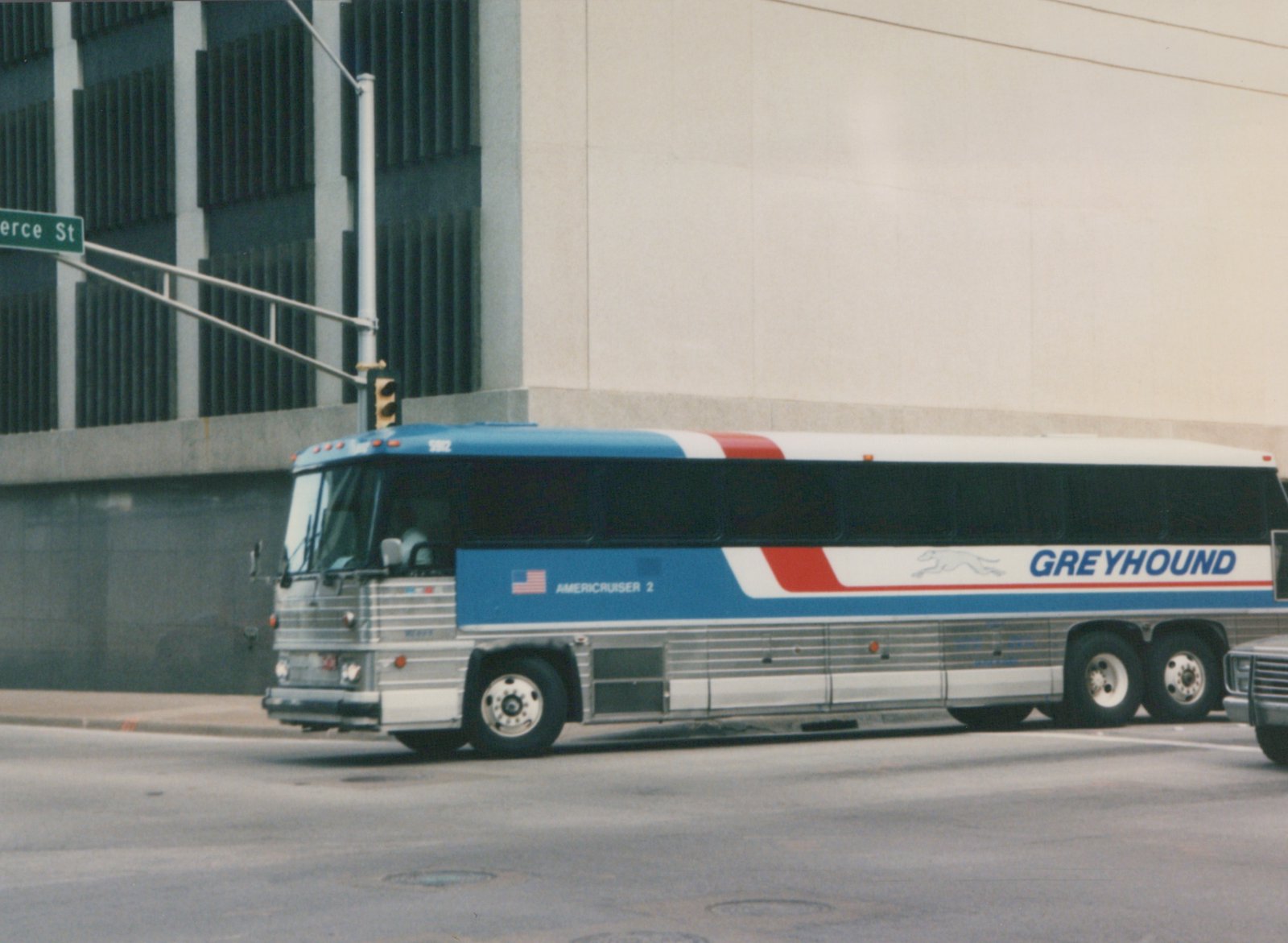A side view of a Greyhound intercity bus, model Flxible Metro, with number 5902 on the windshield, driving on a city street with a green street sign for 'erce St' visible.
