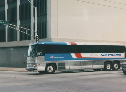 A side view of a Greyhound intercity bus, model Flxible Metro, with number 5902 on the windshield, driving on a city street with a green street sign for 'erce St' visible.