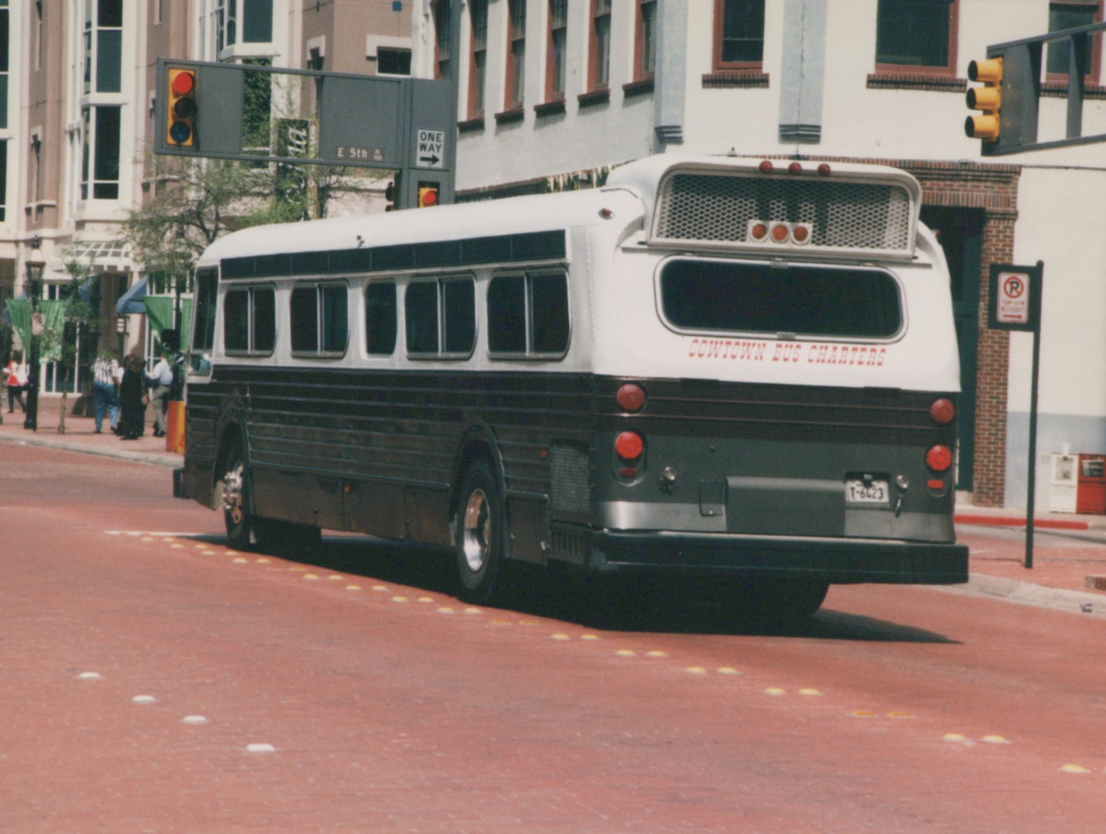 A white and black motor bus with 'Cowtown Bus Charters' lettering on the rear is parked on a city street next to a sidewalk where several people are standing.