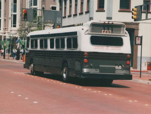 A white and black motor bus with 'Cowtown Bus Charters' lettering on the rear is parked on a city street next to a sidewalk where several people are standing.