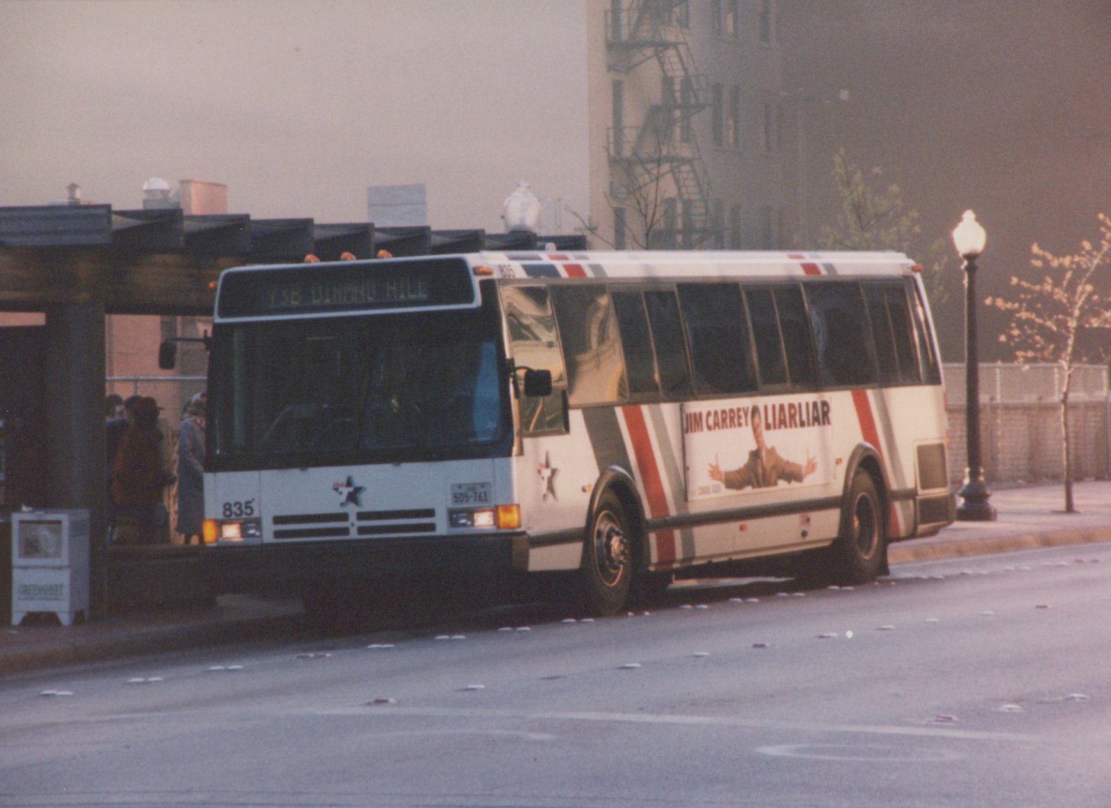 A white Fort Worth Transit bus with red and gray stripes, number 835, shows a 'Jim Carrey Liar Liar' ad on its side as it stops at a street corner with people waiting.