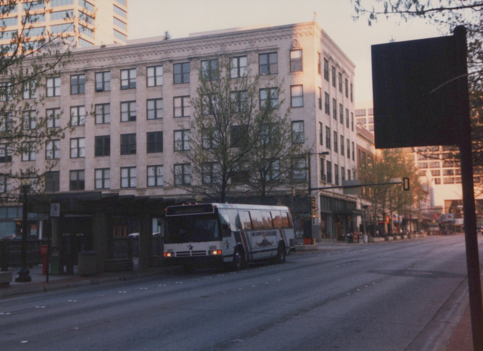 A white city bus with red and blue stripes is parked on an empty street at dusk, with a large building and trees in the background.