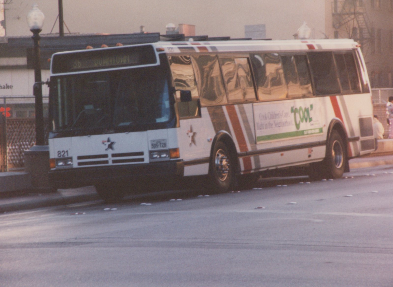 A white motor bus with red and grey stripes and a star logo, displaying route 36 Downtown, is parked on a street with a storefront and two people in the background.