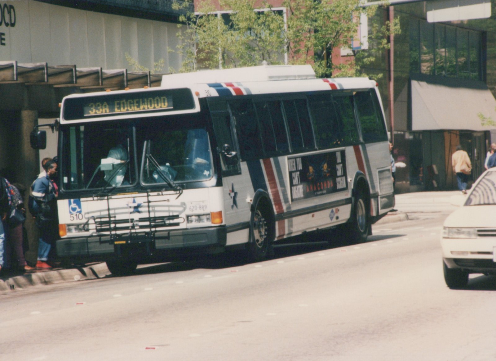 A white, red, and blue striped motor bus numbered 510 is stopped on a city street for passengers, with a movie ad for 'Anaconda' on its side.
