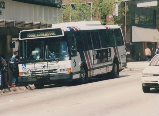 A white, red, and blue striped motor bus numbered 510 is stopped on a city street for passengers, with a movie ad for 'Anaconda' on its side.