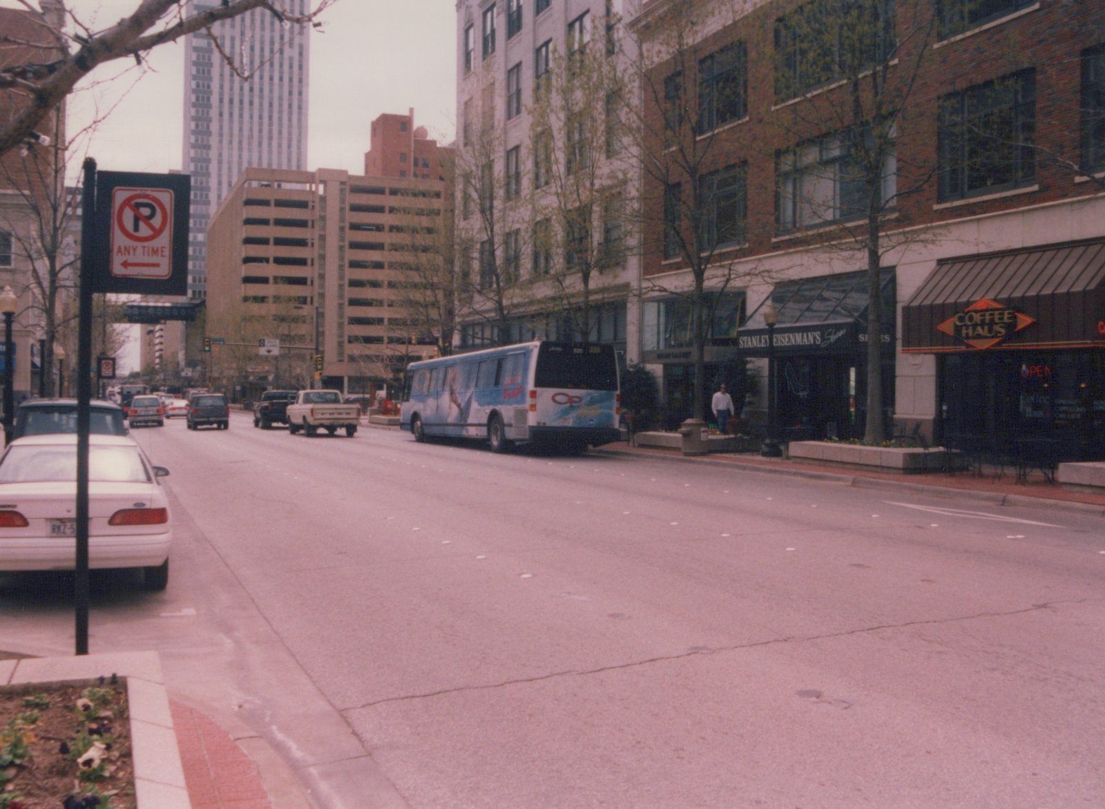 A blue and white bus with an 'OP' logo is parked on a city street, with storefronts including 'Coffee Haus' and 'Stanley Eisenman's' visible nearby, and a few cars and one person on the sidewalk.