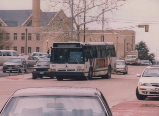 A white transit bus with the number 860 and a lone star logo is driving down a street in Fort Worth, Texas, with several cars and a building in the background.