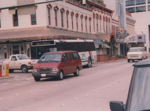 A white city bus with red and blue stripes, number 834, is on a street in Fort Worth, Texas, with a red van in front of it and a movie theater sign visible behind.