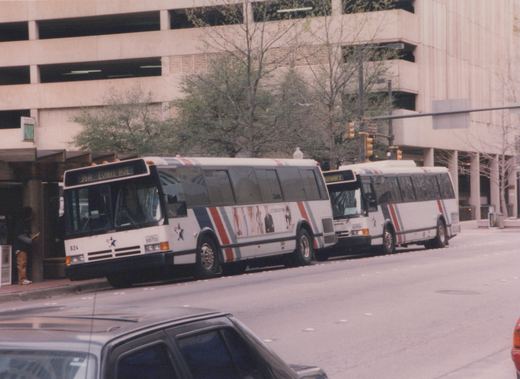 Two white, red, and blue Fort Worth Transit buses on a city street, with a multi-level parking garage in the background.